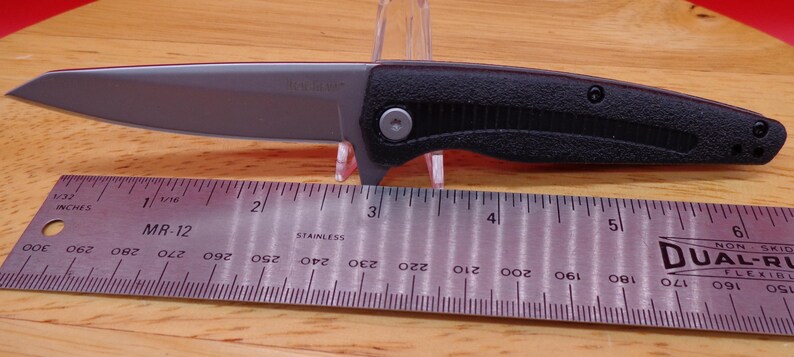 May include: A black and silver folding knife with a partially serrated blade. The knife is shown with a ruler, and the blade appears to be approximately 3 inches long. The handle is black and textured.