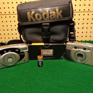 May include: Two silver Kodak Advantix cameras with black carrying case. The camera on the left is a Kodak Advantix 2.0 and the camera on the right is a Kodak Advantix C 400.