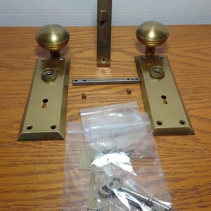 May include: A set of antique brass door hardware, including two door knobs, two plates, a latch, a strike plate, a key, and a bag of screws. The hardware is in good condition and has a beautiful patina.