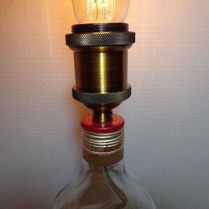 K&ouml;nnte beinhalten: Handgefertigte Lampe aus einer wiederverwendeten Glasflasche. Die Lampe hat eine messingfarbene Fassung und einen roten Flaschenverschluss als Basis. Die Gl&uuml;hbirne ist eine Edison-Gl&uuml;hbirne im Vintage-Stil, die ein warmes Licht ausstrahlt. Ein einzigartiges Wohnaccessoire.
