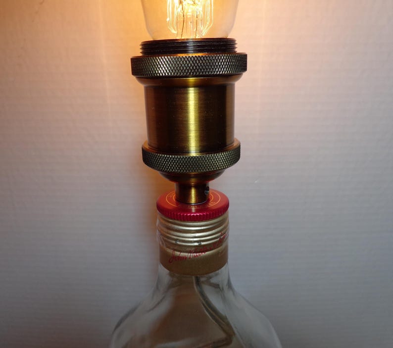 K&ouml;nnte beinhalten: Handgefertigte Lampe aus einer wiederverwendeten Glasflasche. Die Lampe hat eine messingfarbene Fassung und einen roten Flaschenverschluss als Basis. Die Gl&uuml;hbirne ist eine Edison-Gl&uuml;hbirne im Vintage-Stil, die ein warmes Licht ausstrahlt. Ein einzigartiges Wohnaccessoire.
