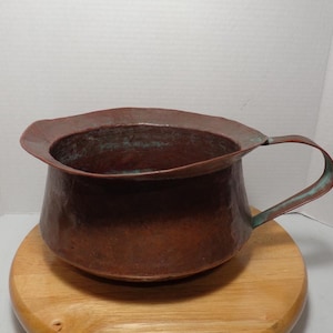 May include: A hammered copper pitcher with a wide, flared rim and a curved handle. The copper has a rich, reddish-brown color with areas of green patina, suggesting age. The pitcher sits on a round wooden surface.
