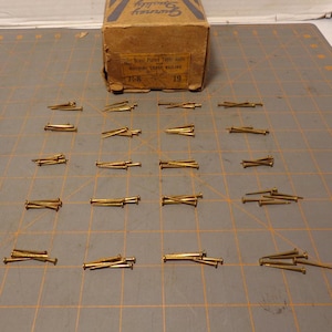 May include: A box of brass plated tack nails for machine made railing. The box is labeled "7-8" and "19". There are 18 piles of nails laid out on a gridded surface.