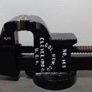 May include: A black metal bench vise with a rusty threaded handle and screw mechanism. The side of the vise features white painted text reading COL. HDW. CO. CLEVELAND U.S.A. NO. 143. The tool has a sturdy base and a sliding jaw assembly.
