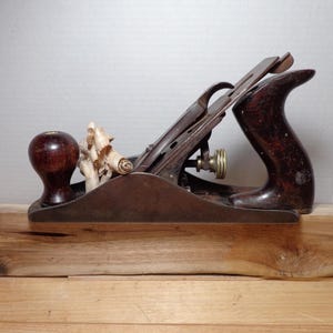 May include: A vintage wood plane with a dark wooden handle and knob. The metal blade and hardware show signs of age. The plane rests on a light-colored wooden block, with wood shavings in the plane's mouth.