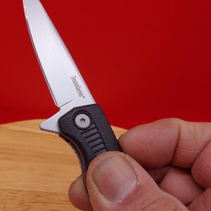 May include: A small, silver folding knife with a black handle. The blade is partially open, revealing the brand name "Kershaw". The knife is held against a red background.