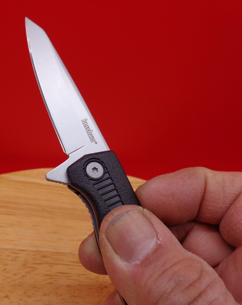 May include: A small, silver folding knife with a black handle. The blade is partially open, revealing the brand name "Kershaw". The knife is held against a red background.