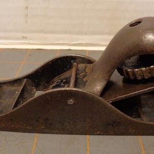 May include: A vintage, well-worn block plane, a woodworking tool, with a dark brown wooden knob and a curved, metal body. The plane has a blade and a lever cap, showing signs of age and use. The tool is designed for smoothing wood surfaces.