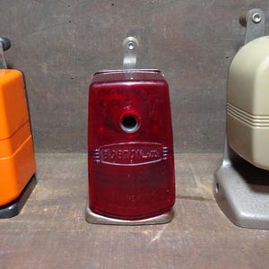 May include: Three vintage pencil sharpeners in orange, red, and beige. The orange sharpener is a cube shape with the text "Hunt Bostonette". The red sharpener is rectangular with the text "Boston-L-M". The beige sharpener has the text "Apsco Sharpeners".