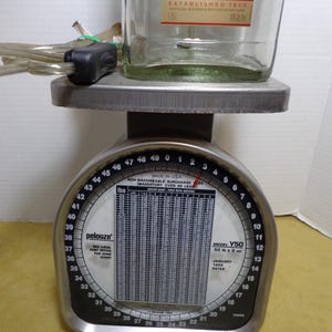 K&ouml;nnte beinhalten: Eine Vintage Pelouze Waage Modell Y50, hergestellt in den USA, mit einer durchsichtigen Glasflasche oben. Die Waage hat ein schwarz-silbernes Zifferblatt mit Messungen in Pfund und Unzen. Die Flasche hat Text.