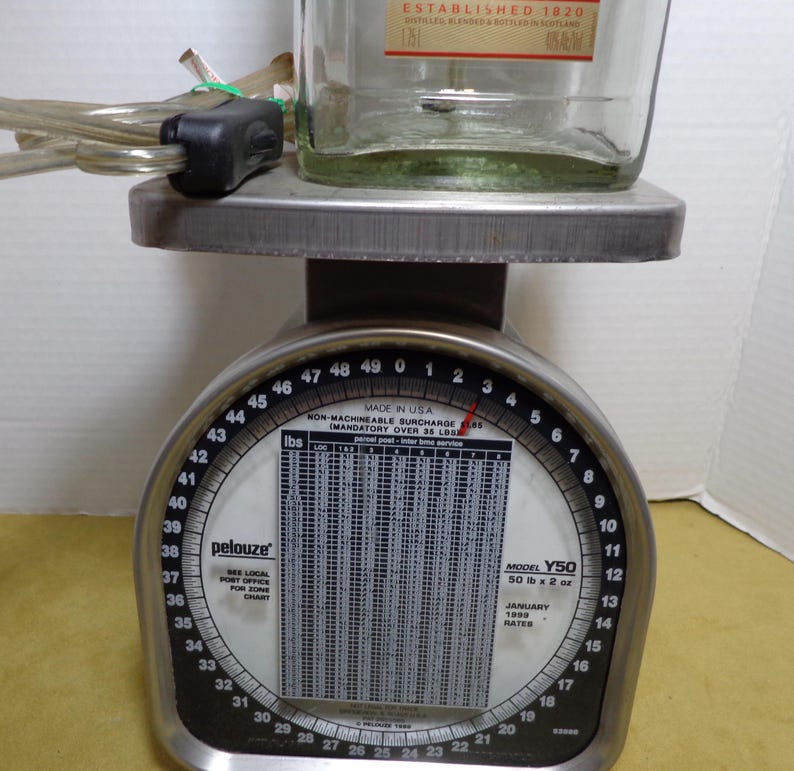 K&ouml;nnte beinhalten: Eine Vintage Pelouze Waage Modell Y50, hergestellt in den USA, mit einer durchsichtigen Glasflasche oben. Die Waage hat ein schwarz-silbernes Zifferblatt mit Messungen in Pfund und Unzen. Die Flasche hat Text.