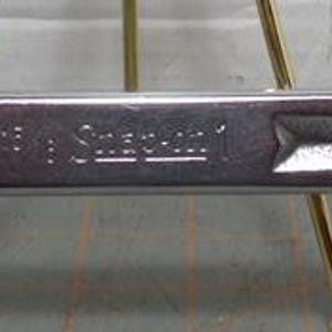 May include: A silver, double-ended wrench with a hexagonal opening on each end. The wrench is made of metal and has a rectangular body with a slightly textured surface. The brand name is visible on the body.