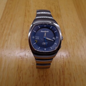 May include: A silver Reebok wristwatch with a blue face and silver hands. The watch has a digital display and the words "Indigo Alarm Wristom" are printed on the face.