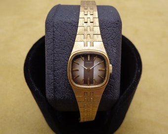 May include: A gold-toned wristwatch with a square face and a brown dial. The watch has a gold-toned metal band and the word "SEIKO" is printed on the face.