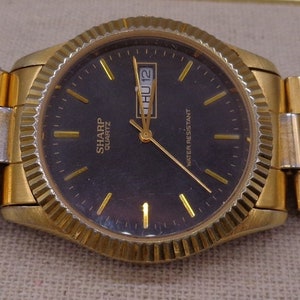 May include: A gold-tone wristwatch with a black face and a gold metal band. The watch face has a date window and the words "Sharp Quartz" printed on it. The watch is water resistant.
