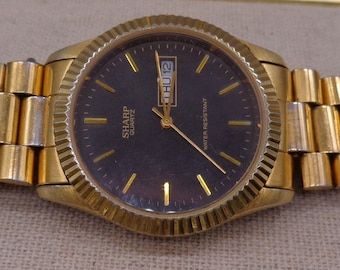 May include: A gold-tone wristwatch with a black face and a gold metal band. The watch face has a date window and the words "Sharp Quartz" printed on it. The watch is water resistant.