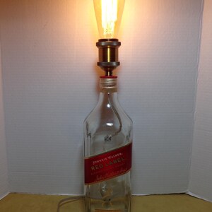 K&ouml;nnte beinhalten: Eine umfunktionierte Lampe aus einer Johnnie Walker Red Label Whiskyflasche. Die transparente Glasflasche hat ein rotes Etikett und ist mit einer messingfarbenen Lampenfassung und einer Gl&uuml;hbirne im Vintage-Stil versehen. Die Lampe enth&auml;lt ein Netzkabel und einen Ein-/Ausschalter.