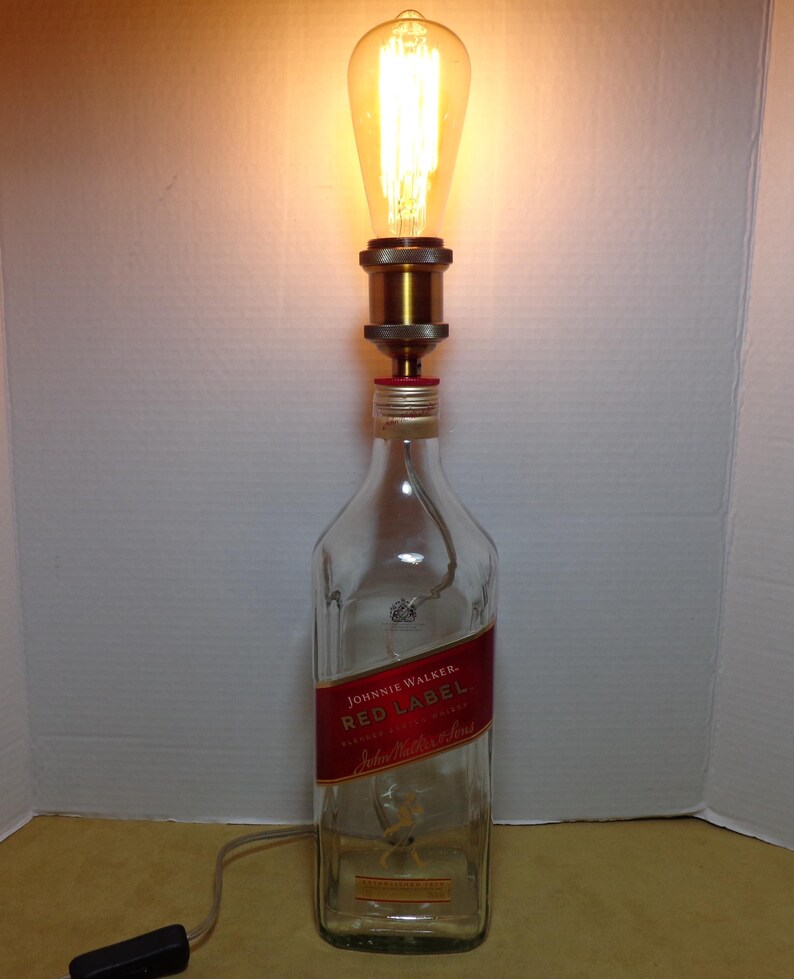 K&ouml;nnte beinhalten: Eine umfunktionierte Lampe aus einer Johnnie Walker Red Label Whiskyflasche. Die transparente Glasflasche hat ein rotes Etikett und ist mit einer messingfarbenen Lampenfassung und einer Gl&uuml;hbirne im Vintage-Stil versehen. Die Lampe enth&auml;lt ein Netzkabel und einen Ein-/Ausschalter.