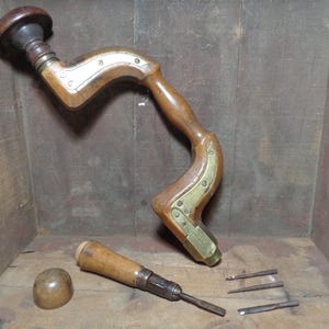 May include: An antique wooden hand drill with brass accents and a dark finish. The drill features a curved handle and a rounded top. Additional tools include a drill bit and a wooden knob. The tools are displayed on a wooden surface.