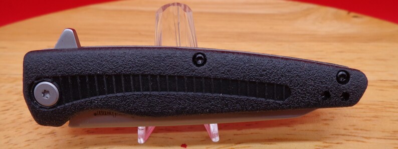 May include: A black folding knife with a textured handle and a silver blade. The knife features a gray thumb stud for easy opening and a pocket clip. The blade is partially visible, and the knife is displayed on a clear stand.