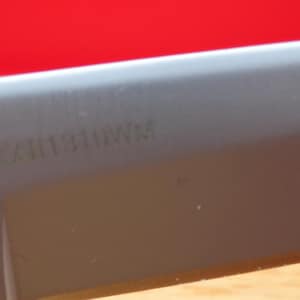 May include: Close-up of a knife blade with the text "KA11310WM" etched into the gray metal. The handle is black and textured. The background is a solid red.