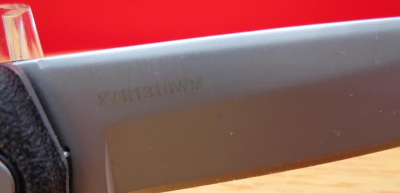 May include: Close-up of a knife blade with the text "KA11310WM" etched into the gray metal. The handle is black and textured. The background is a solid red.