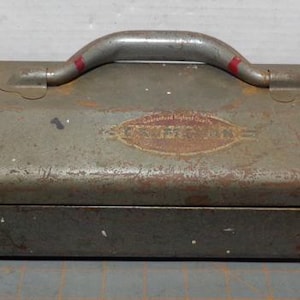 May include: An aged, olive-green metal toolbox with a curved handle and two latches. The toolbox has a faded logo on the front and shows signs of wear and rust. The handle has red accents.