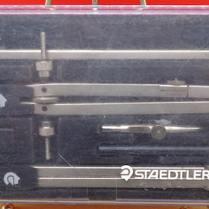 May include: A set of Staedtler Mars drafting tools in a clear plastic case. The set includes a compass, dividers, and other drawing instruments. The tools are silver-colored and arranged in a black foam insert. The case has a red and yellow border.