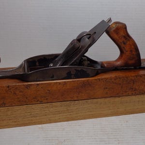 May include: A vintage wooden hand plane with a dark metal blade and a wooden handle. The plane is set on a wooden base. The handle and knob are made of a lighter-colored wood. The plane is designed for woodworking and carpentry.