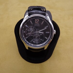 May include: A silver Guess watch with a black leather band and a black face. The watch has multiple subdials and a date window.