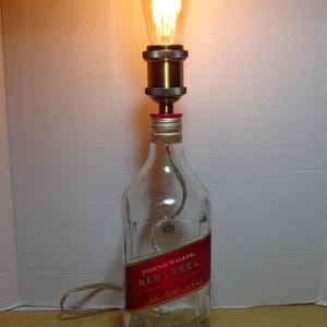 K&ouml;nnte beinhalten: Eine umfunktionierte Lampe aus einer Johnnie Walker Red Label Whiskyflasche. Die transparente Glasflasche hat ein rotes Etikett mit goldfarbenen Buchstaben. Eine dekorative Gl&uuml;hbirne sitzt oben auf der Flasche, mit einer messingfarbenen Metallfassung und einem sichtbaren Netzkabel.