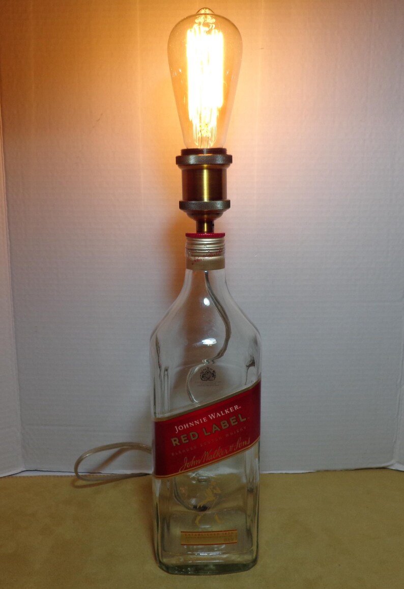 K&ouml;nnte beinhalten: Eine umfunktionierte Lampe aus einer Johnnie Walker Red Label Whiskyflasche. Die transparente Glasflasche hat ein rotes Etikett mit goldfarbenen Buchstaben. Eine dekorative Gl&uuml;hbirne sitzt oben auf der Flasche, mit einer messingfarbenen Metallfassung und einem sichtbaren Netzkabel.