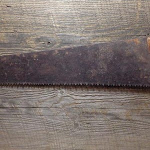 May include: An antique hand saw with a dark, aged blade and a wooden handle. The saw blade is long and has many teeth. The handle is a light brown color and has a curved design. The saw is mounted on a wooden surface.