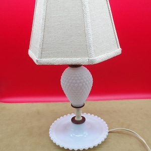 May include: White milk glass table lamp with a textured, bulbous base and a white linen lampshade. The lamp has a round, beaded base.