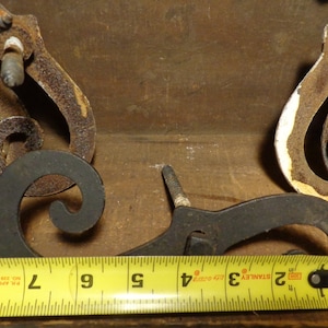 May include: Three antique metal door hinges with ornate scrollwork. The hinges are rusty and have a black finish. A yellow measuring tape is in the foreground, showing measurements in inches.