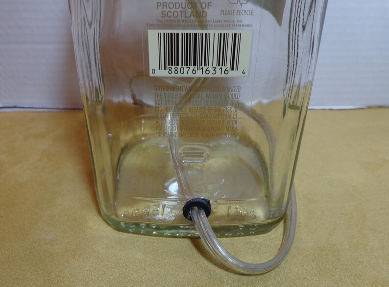 K&ouml;nnte beinhalten: Eine klare Glasflasche mit einem durch den Boden eingef&uuml;hrten, durchsichtigen Schlauch. Die Flasche hat ein Etikett mit Text und einem Barcode. Der Schlauch wird von einer schwarzen Gummit&uuml;lle gehalten. Die Flasche steht auf einer gelben Oberfl&auml;che.