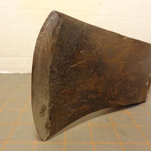 May include: Close-up of a vintage axe head. The axe head is made of metal with a rusty brown color and a curved blade. The blade has a silver edge. The axe head is likely used for chopping wood or other similar tasks.