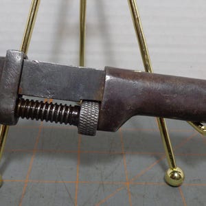 May include: An antique adjustable wrench with a dark, weathered appearance. The wrench features a screw adjustment mechanism and a textured handle. The metal tool is displayed on a gold-colored stand, showcasing its vintage design.