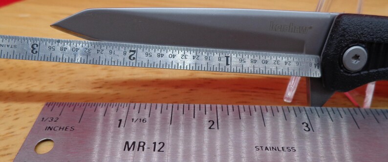 May include: A silver folding knife with a black handle is measured with a ruler. The blade is approximately 3 inches long. The knife is on a wooden surface.
