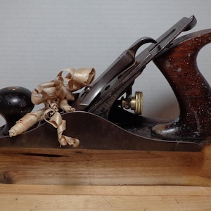 May include: A vintage hand plane with a wooden base and a dark metal blade. The plane has a wooden handle and knob, with wood shavings resting on top. The tool is designed for woodworking.