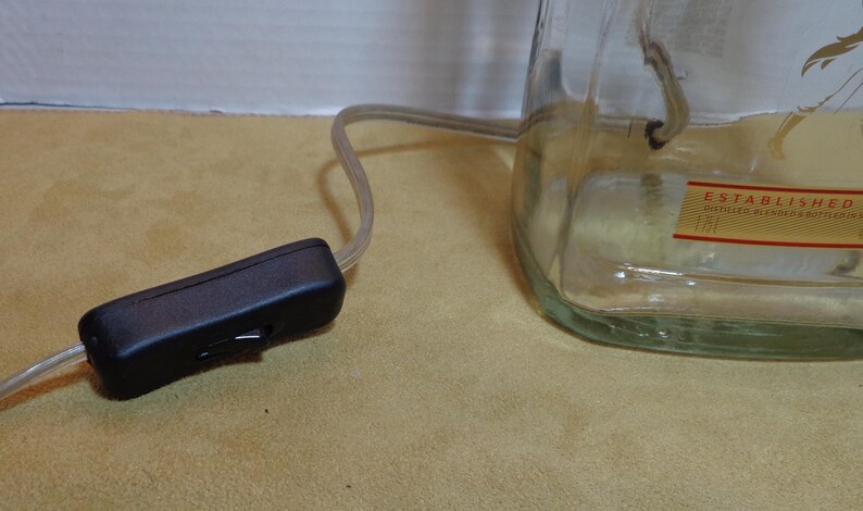 K&ouml;nnte beinhalten: Eine klare Glasflasche, die als Lampensockel wiederverwendet wurde, mit einer Gl&uuml;hbirne im Inneren. Ein schwarzer Schnurschalter ist mit einem transparenten Elektrokabel verbunden. Die Flasche hat ein Etikett mit der Aufschrift "ESTABLISHED".