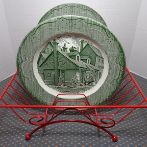 May include: A set of green and white decorative plates featuring a village scene, displayed in a red metal plate rack. The plates have a detailed design and are stacked in the rack, which has a curved base and a handle.