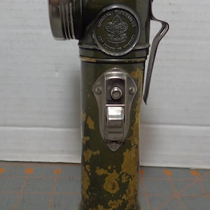 May include: Vintage olive green flashlight with a silver bezel and handle. The flashlight has a circular emblem that reads "Official Boy Scouts of America." The body shows signs of wear with yellowed paint. A switch is located on the front.