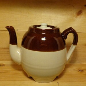 May include: A brown and white ceramic teapot with a lid. The teapot has a rounded body and a spout on the side. The handle is on the opposite side of the spout.
