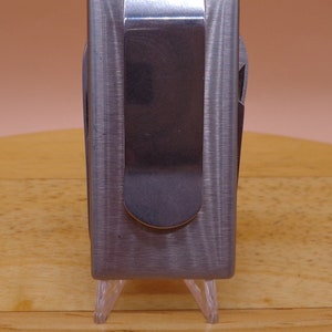 May include: A silver metal money clip with a brushed finish. The clip is closed and has a smooth, rounded edge.