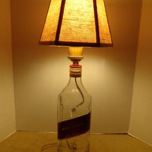 K&ouml;nnte beinhalten: Eine einzigartige Tischlampe aus einer wiederverwendeten Flasche. Die Lampe hat einen rechteckigen Lampenschirm aus braunem Stoff und einen Sockel aus durchsichtigem Glas mit einem roten Etikett. Die Lampe ist eingesteckt und einsatzbereit.