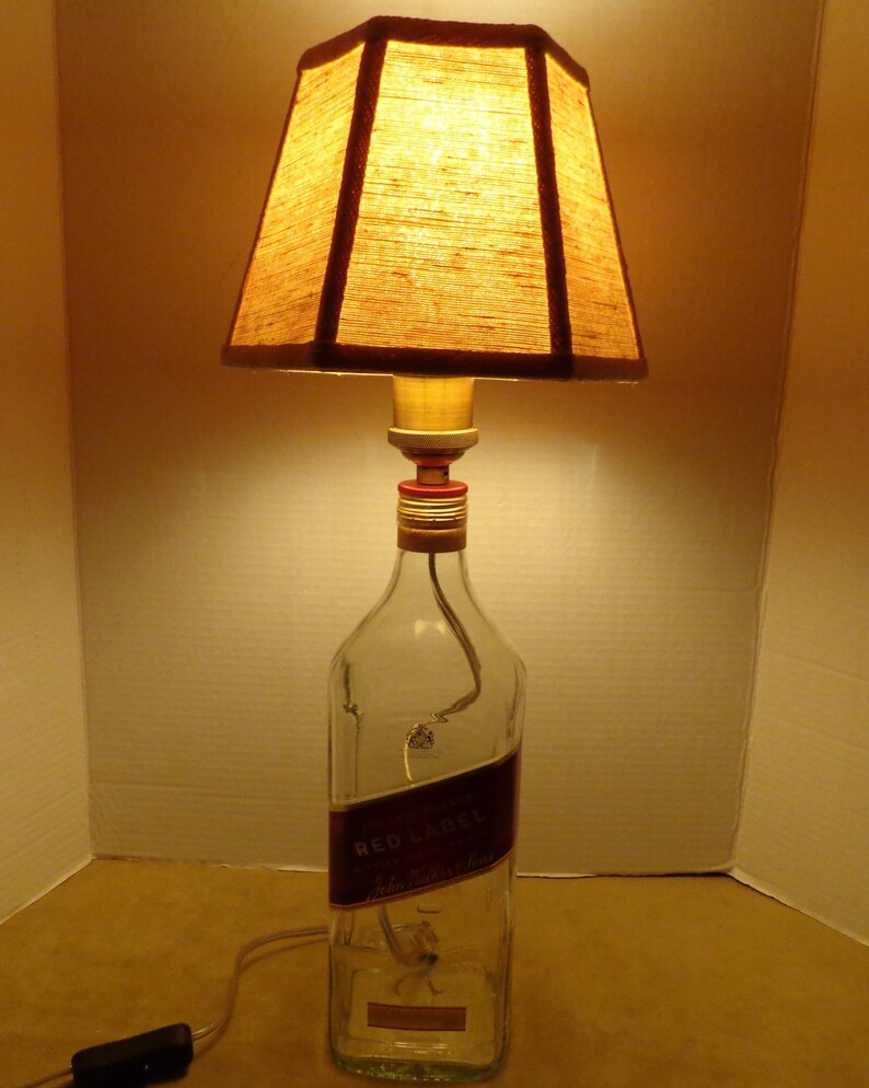 K&ouml;nnte beinhalten: Eine einzigartige Tischlampe aus einer wiederverwendeten Flasche. Die Lampe hat einen rechteckigen Lampenschirm aus braunem Stoff und einen Sockel aus durchsichtigem Glas mit einem roten Etikett. Die Lampe ist eingesteckt und einsatzbereit.