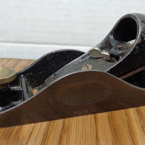 May include: A vintage hand plane, likely for woodworking. The plane is primarily black with a wooden base and brass adjustment knobs. The blade is visible, and the tool is resting on a wooden surface. The text "No. 9 1/4" is visible on the base.