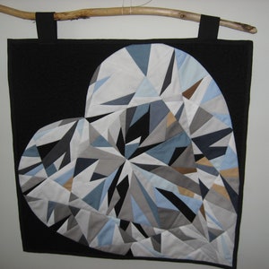 Puede incluir: Una pared colgante de tela negra con un gran diseño de corazón en forma de diamante. El corazón está hecho de varios tonos de azul, gris y blanco, piezas de tela cosidas juntas.