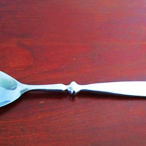 May include: A silver spoon with a rounded bowl and a simple, elegant design. The spoon is lying on a dark red wooden surface.